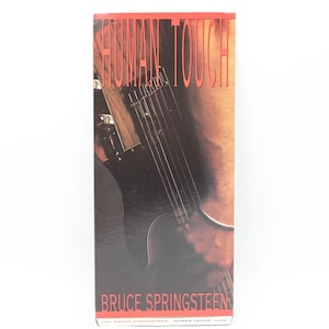 May include: A rectangular box with the words "HUMAN TOUCH" in red block letters at the top. Below, a black electric guitar is visible, with the name "BRUCE SPRINGSTEEN" at the bottom. The background is a mix of brown and black.