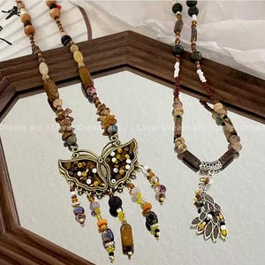 Peut inclure: Deux colliers de perles avec des pendentifs décoratifs. L'un des colliers présente un pendentif en forme de papillon avec des perles brunes, dorées et blanches. L'autre collier a un pendentif en forme de feuille avec des perles brunes, blanches et vertes.