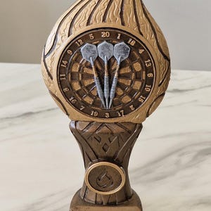 May include: A gold-coloured trophy shaped like a flame with a dartboard and three darts in the centre. The base of the trophy is brown and has a gold-coloured medallion with a flame design.