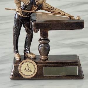 Könnte beinhalten: Bronze-farbene Billardspieler-Figur in Schusspose, mit Queue und Billardtisch. Die Figur steht auf einem rechteckigen Sockel mit einem kreisförmigen Emblem von Billardkugeln und einer leeren Plakette.