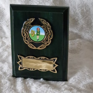 Puede incluir: Una placa de madera verde con un diseño de nudo celta dorado y una imagen redonda y colorida de una mujer con un vestido verde sosteniendo un arpa. La placa tiene un espacio rectangular dorado para grabar.