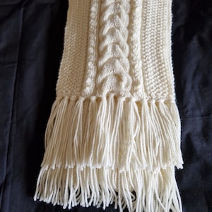 May include: A white knitted scarf with a cable knit pattern and a long fringe.