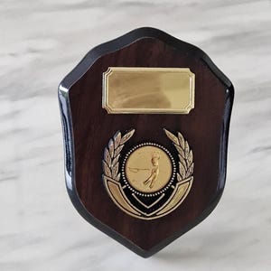 Könnte beinhalten: Golf-Auszeichnungstafel aus Holz mit dunkelbraunem Finish. Die Tafel zeigt ein goldfarbenes Emblem eines Golfspielers, umgeben von einem Lorbeerkranz. Oben befindet sich eine goldene Platte zum Gravieren.