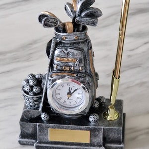Könnte beinhalten: Eine silberne Golftasche mit einer Uhr und einem Stifthalter. Die Tasche ist mit Golfschlägern und Golfbällen gefüllt. Das Zifferblatt der Uhr ist weiß mit schwarzen Zahlen und Zeigern. Der Stifthalter ist golden und hat einen schwarzen Stift.