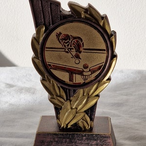 May include: A gold and brown trophy with a laurel wreath design. The centre of the trophy features a gold circle with a depiction of a person playing snooker.
