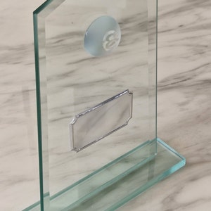 Puede incluir: Una placa de premio de vidrio transparente con una placa plateada para grabar. La placa tiene un borde biselado y un pequeño círculo transparente con el número 3 en el centro.