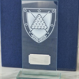 Puede incluir: Trofeo de vidrio transparente con forma rectangular y base. El trofeo presenta un gráfico blanco de un juego de billar, tacos y estrellas dentro de un escudo. Una placa de identificación plateada en la parte inferior.