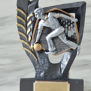 May include: A bowling trophy with a silver bowler in mid-action, about to release the ball. The trophy has a black base with a gold nameplate, and a shield-shaped frame with a gold border. A gold laurel leaf design is on the side.