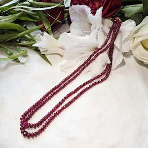 Könnte beinhalten: Eine dreireihige Halskette aus kleinen, facettierten, tiefroten Perlen. Die Halskette liegt auf einer weißen Oberfläche, mit weißen und roten Blumen im Hintergrund. Die Perlen haben einen leichten Glanz.