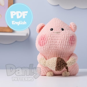 May include: A handmade pink crochet pig with rosy cheeks, holding a brown and beige acorn. The pig has small black eyes and ears. The image includes the text "PDF English" in a blue circle and "Danty PATTERNS" in white.