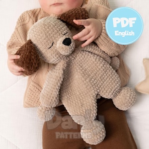 May include: A soft, beige dog-shaped plush toy with brown ears and a black nose. The toy is held by a baby wearing a beige sweater and brown pants. The image includes the text "PDF English" in a blue circle.