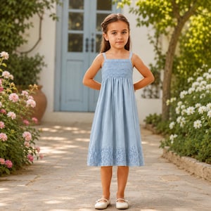 May include: A pale blue dress with a smocked bodice and thin straps. The dress has a detailed, eyelet lace hem. The girl is wearing white flats. The background includes a blue door, flowers, and a stone pathway.