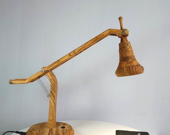 Könnte beinhalten: Eine Holz-Schreibtischlampe mit natürlichem Finish. Die Lampe hat einen verstellbaren Arm und einen konischen Lampenschirm. Der runde Sockel ist mit einem Netzkabel verbunden. Ein Notizbuch und ein Stift liegen auf dem Tisch.