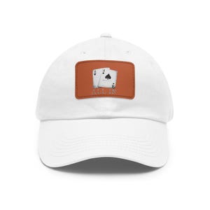 Puede incluir: Gorra de béisbol blanca con un parche de cuero marrón que presenta dos cartas de juego, un as de picas y un rey de picas, con el texto "ALL IN" debajo de las cartas.