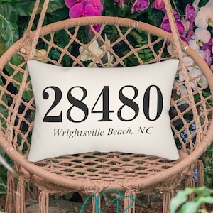 May include: A white pillow with black text that reads "28480 Wrightsville Beach, NC" is sitting on a woven hanging chair. The chair is surrounded by pink and white orchids.