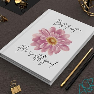 May include: A white journal with a pink flower and the text "But if not, He is still good" printed on the cover.