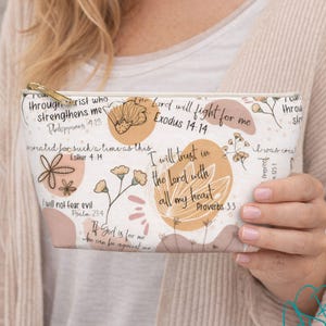May include: A white cosmetic bag with a gold zipper, featuring floral and script designs. The bag includes inspirational quotes and phrases in black, brown, and peach colors. The bag is held by a person.