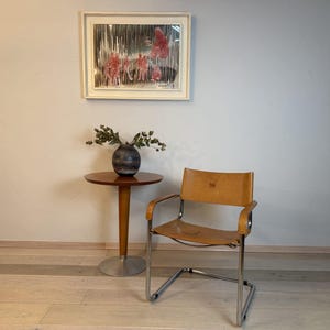 Könnte beinhalten: Innenaufnahme eines Raumes mit einem runden Beistelltisch aus Holz, einer dunklen Vase mit Grün und einem gerahmten Kunstwerk. Rechts befindet sich ein Vintage-Stuhl mit hellbraunem Ledersitz und Chromrahmen. Der Raum hat eine neutrale Farbpalette.