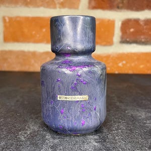 Könnte beinhalten: Keramikvase mit dunkelblauer und violetter gesprenkelter Glasur. Die Vase hat einen zylindrischen Körper und einen kurzen, ausgestellten Hals. Der Text "OTTOKERAMIK" befindet sich auf einem kleinen Etikett auf der Vase.