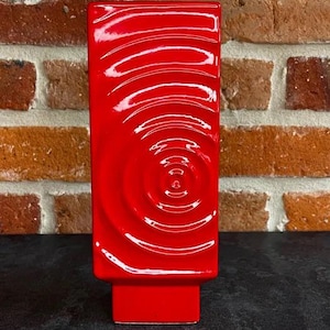 Può includere: Un vaso rettangolare rosso vivo con un design a cerchi concentrici strutturati. Il vaso ha una finitura lucida e una base rettangolare. Lo sfondo presenta un muro di mattoni.