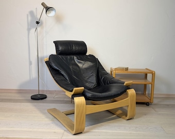 May include: A mid-century modern lounge chair with a black leather seat and headrest, set on a curved, light-colored wooden frame. A matching side table and a silver floor lamp are also in the room.