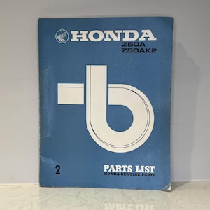 May include: A blue Honda Z50A Z50AK2 parts list manual with white text and a large white graphic design. The cover features the Honda logo, the model numbers, the number 2, and the text PARTS LIST HONDA GENUINE PARTS printed in white.