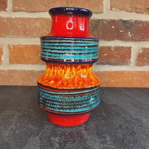 Puede incluir: Un jarrón de cerámica vibrante con un diseño único en capas. El jarrón presenta una base y una parte superior rojas, con una sección central de rayas turquesas y negras. La sección media hace la transición de naranja a rojo, creando un contraste visual llamativo.