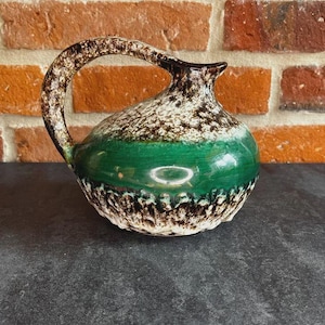 Może przedstawiać: Ceramiczny dzban z zakrzywionym uchwytem. Dzban ma zielony pas wokół środka, z nakrapianym brązowo-białym wzorem powyżej i poniżej. Obręcz i uchwyt są brązowe.