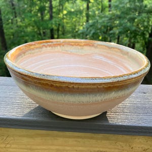 Puede incluir: Un cuenco de cerámica hecho a mano con un interior rosa claro y un borde degradado marrón, beige y azul. El cuenco está sobre una superficie de madera con un fondo verde borroso de árboles.