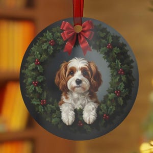 May include: Round Christmas ornament featuring a Cavalier King Charles Spaniel dog inside a wreath of holly and red berries. A red ribbon and bow are at the top. The ornament is hanging from a red ribbon.