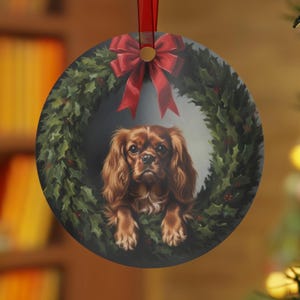 May include: A round Christmas ornament featuring a Cavalier King Charles Spaniel dog within a wreath of green holly with red berries. A red bow adorns the top, and a red ribbon is attached for hanging. The ornament has a festive holiday theme.