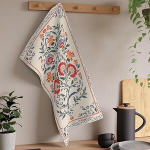 May include: Cream-colored kitchen towel with a floral design in red, blue, and orange. The towel is hanging on a wooden rack. A black teapot, plates, and cups are on a wooden countertop.
