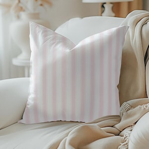 May include: A decorative throw pillow with vertical pink and white stripes. The pillow is placed on a white sofa, with a beige blanket draped over the arm. A lamp and vase are visible in the background.
