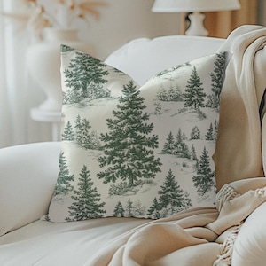 May include: A decorative throw pillow with a forest scene print. The pillow features a repeating pattern of green pine trees and other foliage against a cream-colored background. The pillow is placed on a white armchair.