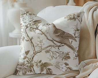 May include: A decorative throw pillow with a bird and floral design in shades of brown and cream. The pillow is placed on a white armchair, with a cream-colored blanket draped over the arm. A lamp and vase are visible in the background.