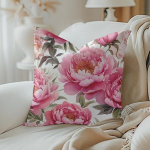 May include: A decorative throw pillow featuring a watercolor floral design of pink peonies and green leaves on a white background. The pillow is placed on a white sofa with a beige throw blanket.