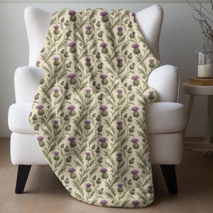 Op de afbeelding: Een zachte plaid met een herhalend botanisch patroon van paarse distelbloemen met groene stengels en bladeren op een crèmekleurige achtergrond. De stof vertoont een consistent bloemmotief over het gehele zichtbare oppervlak.