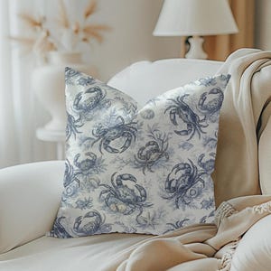 May include: A decorative pillow with a blue and white crab pattern. The pillow features a repeating design of crabs, seaweed, and seashells. The pillow is placed on a white sofa with a beige throw blanket.