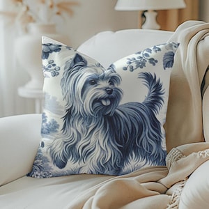 May include: A white cushion with a blue and white floral pattern featuring a Yorkshire Terrier dog. The cushion is sitting on a white sofa with a beige throw blanket.