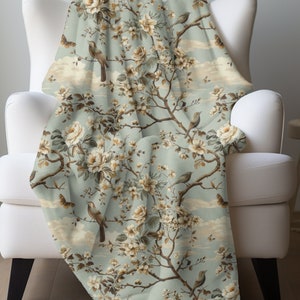 May include: A soft, blue throw blanket with a floral pattern featuring delicate white flowers and brown birds. The blanket has a subtle cloud-like pattern in the background.