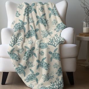 May include: A soft, cream-colored throw blanket with a repeating pattern of blue sea turtles and coral. The blanket is draped over a white armchair.