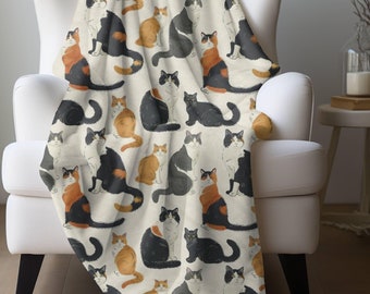 May include: A cream-colored throw blanket with a repeating pattern of black, orange, and white cats. The blanket is draped over a white armchair.
