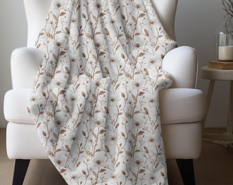 May include: A white throw blanket with a brown floral pattern featuring delicate branches and small blossoms. The blanket is draped over a white armchair.