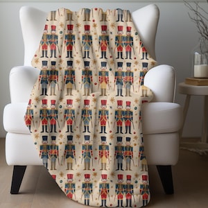 May include: A cream-colored throw blanket with a repeating pattern of toy soldiers in red, blue, and gold. The soldiers are holding rifles and have stars on their hats. The background is a cream color with gold stars.