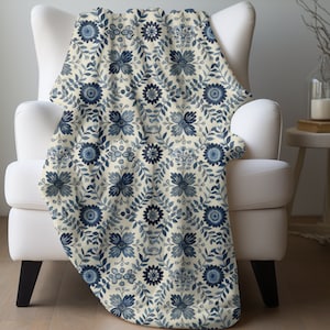 May include: A cream-colored throw blanket with a blue floral pattern. The blanket is draped over a white armchair.