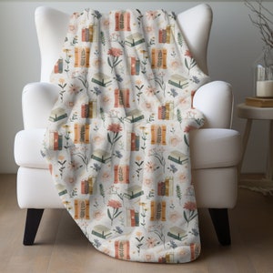 May include: A white throw blanket with a repeating pattern of books and flowers in shades of pink, orange, and green. The blanket is draped over a white armchair.