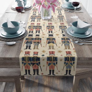 May include: A beige table runner with a repeating pattern of toy soldiers in red, blue, and black uniforms. The runner is decorated with small star and dot accents. The table is set with plates, bowls, and wine glasses.