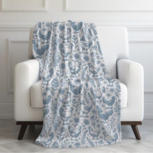 May include: A blue and white throw blanket draped over a white armchair. The blanket features a repeating pattern of chickens, flowers, and foliage. The design is in a soft, muted blue against a white background, creating a cosy and inviting aesthetic.