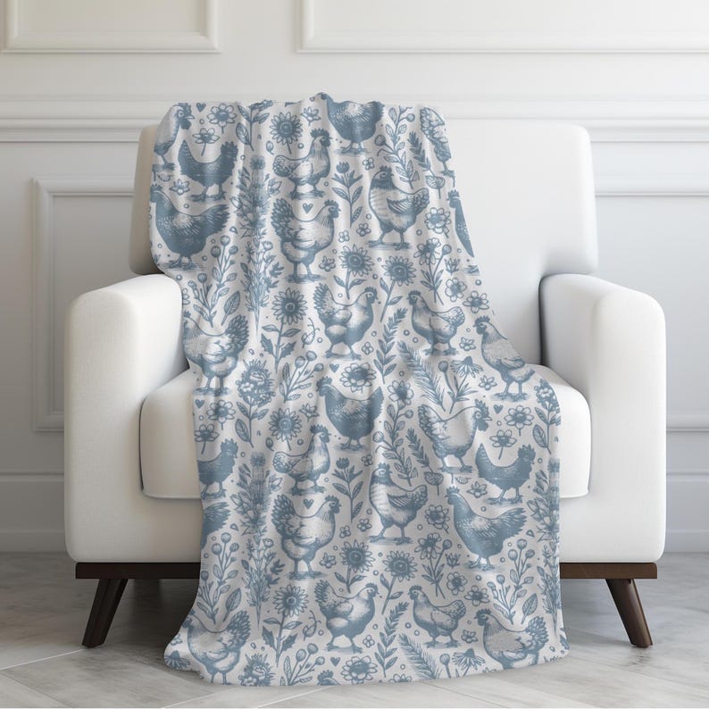 May include: A blue and white throw blanket draped over a white armchair. The blanket features a repeating pattern of chickens, flowers, and foliage. The design is in a soft, muted blue against a white background, creating a cosy and inviting aesthetic.