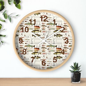 May include: Round wall clock with a light wood frame. The clock face features a cream-colored background with a pattern of various fish and fishing lures. Brown numerals mark the hours, and the clock hands are white.